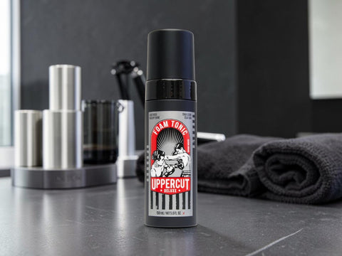 A bottle of Uppercut Team Tonic hair product standing on a dark countertop, with a blurred modern grooming setup in the background including metallic containers and a folded dark towel.